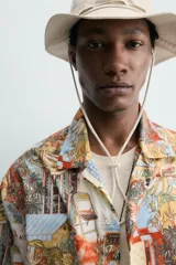 Camisa de corte relajado confeccionada en tejido ligero de algodón, con cuello bowling y manga corta. Presenta un estampado integral con motivos urbanos y tropicales en tonos cálidos.