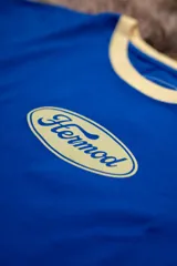 Remera corta de manga corta, color azul vibrante, con cuello redondo y ribetes en color crema. Presenta un logo ovalado centrado con el texto "Hermod" en tipografía cursiva.