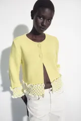 Chaqueta de punto color amarillo claro, con cuello redondo y mangas por debajo del codo. Presenta detalles de acabado en tejido crochet blanco en el ruedo y los puños, con cierre frontal de botones forrados al tono.