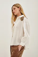 Camisa blanca de algodón con cuello camisero y mangas largas abullonadas con puños ajustados. Presenta detalles bordados en los hombros con motivos vegetales y aplicaciones brillantes.