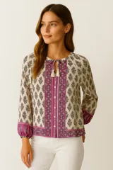 Blusa color crema con estampado de flores y motivos geométricos en tonos rosados y morados. Tiene cuello redondo con lazo y borlas, mangas largas abullonadas con puños elásticos y cierre frontal con botones.