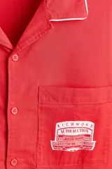 Camisa de manga corta color rojo con cuello abierto en contraste blanco, cierre frontal de botones y un bolsillo en el pecho con estampa gráfica.