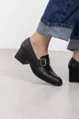 Mocasín de cuero marrón con tacón ancho efecto madera, puntera cuadrada y tira lateral con hebilla metálica y detalle texturizado.