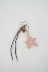 Llavero con mosquetón en forma de corazón que incluye una tira de cuero sintético rosado con un dije de estrella de mar decorado con tachas doradas, un cordón gris con detalles reflectivos y un dije en forma de caracol iridiscente.