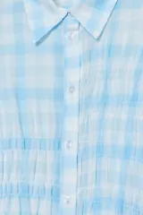 Camisa de manga larga con estampado de cuadros vichy en tonos celeste y blanco. Presenta cuello clásico, cierre frontal con botones y detalle de fruncido en la parte central delantera.