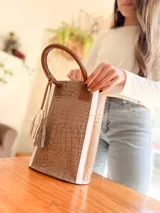 Bolso matero de cuero color taupe con textura croco y detalles en cuero color marrón. Tiene doble asa de mano y un dije de borla.