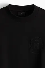 Buzo negro de tejido pesado con interior cepillado suave, cuello redondo y corte boxy. Presenta un diseño estampado en relieve tono sobre tono en el pecho.