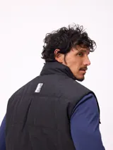 Chaleco acolchado color negro con cierre frontal de cremallera y botones a presión. Presenta cuello alto, bolsillos frontales con solapa y un pequeño parche con logo en el pecho.