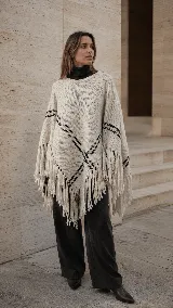 Poncho tejido en lana merino de diseño asimétrico, color crudo con detalles de líneas geométricas en negro y terminaciones con flecos largos en todo el borde.