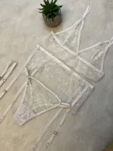 Conjunto de lencería de encaje blanco compuesto por un bralette triangular, una tanga con tiras laterales y un portaligas integrado. El diseño presenta detalles de encaje floral y breteles regulables.