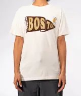 Remera color crudo con cuello redondo y manga corta. Estampado frontal de una banderín marrón con la palabra "BOSTON" en letras doradas con efecto glitter. Incluye dos pines decorativos prendidos en la zona superior derecha del pecho.