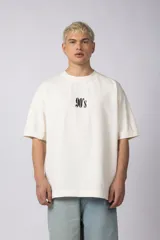 Remera oversize de algodón 100% premium, color blanco hueso, con bordado frontal "90's" en negro y un parche trasero con una imagen estilo videoclip de Nirvana.