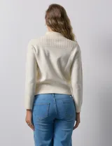 Cardigan de tejido punto color crema, con cuello tipo solapa, cierre frontal completo con tirador circular metálico y dos bolsillos de parche simulados en el frente. Presenta detalles de canalé en los hombros.