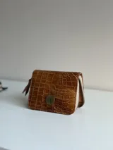 Billetera pequeña de cuero color tostado con textura símil cocodrilo, cierre perimetral de cremallera y detalle de aplique metálico circular en el frente.
