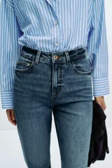Jeans de mujer, tiro alto, corte Mom Fit con pierna slim y largo tobillero. Denim azul medio con efecto desgastado y lavado. Presenta cinco bolsillos y cierre frontal con cremallera y botón.