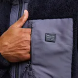 Canguro de sherpa con cierre frontal completo, cuello alto y bolsillo en el pecho con cierre. Presenta una combinación de texturas en color negro y gris oscuro, con diseño funcional y térmico.