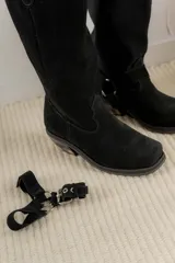 Bota estilo tejana de caña alta, confeccionada en cuero marrón con acabado texturizado. Presenta punta cuadrada, taco bajo y una correa con hebilla metálica ajustable y estribo desmontable en la zona del tobillo.