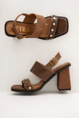 Sandalias de cuero marrón, con taco ancho, faja ancha en el empeine, tira frontal con tachas metálicas y punta cuadrada. Cuentan con ajuste mediante hebilla al tobillo.