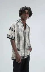 Camisa de manga corta color beige, tejida con punto abierto (símil crochet) y detalles en negro. Tiene cuello solapa, cierre frontal con botones y calce oversize.