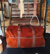 Bolso de mano grande tipo duffel, confeccionado en gamuza marrón con detalles de cuero oscuro en las asas, base y correas verticales de refuerzo. Cuenta con amplios bolsillos frontales abiertos y cierre superior con cremallera.