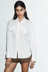 Camisa de popelín de algodón con cuello subido y detalle de aplicación tipo pañuelo anudado al cuello. Presenta manga larga con puños clásicos y cierre frontal mediante botones.