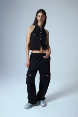 Chaleco de denim negro con escote halter, cuello camisero y cierre frontal con botones metálicos. Presenta dos bolsillos frontales con solapa y diseño sin mangas.
