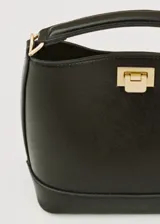 Bolso de mano tipo bucket color negro, con estructura rígida y asa corta superior. Presenta un cierre metálico frontal en color dorado y detalles de herrajes a juego.
