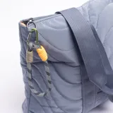Cartera tipo tote bag de diseño acolchado con costuras curvas, color azul grisáceo. Cuenta con doble asa de hombro y un detalle de cordón decorativo con terminales en amarillo y gris en el lateral.