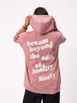 Canguro oversize color rosado con capucha, confeccionado en tejido de algodón y poliéster. Presenta un estampado gráfico en la espalda con la frase 'Dream beyond the edge of reality' y el logo de la marca Rusty.