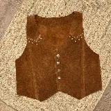 Chaleco de cuero gamuzado color marrón, con cierre frontal de botones metálicos y detalle de tachas decorativas en la zona de los hombros.