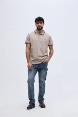 Chomba de piqué color beige, de manga corta, con cuello y puños tejidos a contratono con líneas oscuras. Cuenta con cartera de tres botones y logo bordado en el pecho.