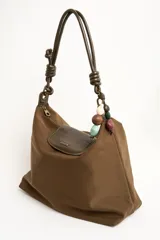 Cartera tipo shopper color marrón, con solapa frontal decorativa en tono más oscuro y tiras con nudos. Incluye herrajes dorados y un charm colgante con cuentas de madera.