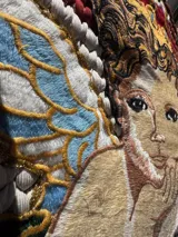 Bolso tejido a mano con hilo de trapillo, eco-cuero y textiles, con un diseño central de querubín coronado y alas, sobre un fondo de franjas rojas, blancas y azules. Posee asas cortas tejidas en color celeste claro.