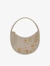 Cartera hobo redonda color crema, con asa de hombro y cierre de cremallera. Decorada con tachas doradas de diferentes tamaños en el frente.