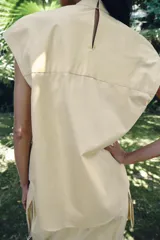Camisa sin mangas confeccionada en algodón, con cuello redondo y detalle de lazada frontal. Presenta un diseño de corte holgado con bajo redondeado y cierre posterior mediante abertura y botones.