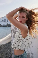 Blusa sin mangas blanca con estampado de lunares negros, cuello alto drapeado y nudo frontal en el bajo.