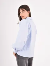 Camisa de manga larga en color celeste, con cuello clásico y cierre frontal de botones. Presenta un diseño con mangas abullonadas y puños ajustados.