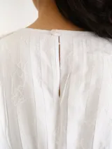 Blusa blanca de manga corta y cuello redondo, con diseño de rayas verticales texturizadas y detalles sutiles de flecos.