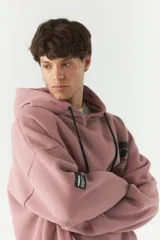 Canguro de felpa color rosa con capucha y cordones negros. Presenta un estampado con texto y figuras en el pecho y un parche con texto en la manga izquierda.