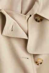 Gabardina de color beige claro, largo hasta la rodilla, de doble botonadura con botones marrones. Presenta solapas clásicas, hombreras, cinturón de tela extraíble con hebilla oscura y bolsillos insertados.