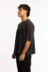 Remera de corte boxy fit, color negro con efecto lavado, de manga corta y cuello redondo. Presenta un logo bordado en el centro del pecho dentro de un óvalo.