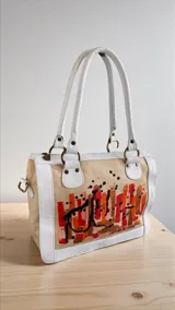 Cartera Diri de cuero natural uruguayo, en tonos marfil y arena, con intervención artística pintada a mano en el frente con pinceladas en rojos, terracotas y dorados. Cuenta con estructura firme, herrajes metálicos en acabado oro viejo, cierres, manijas ergonómicas y correa regulable desmontable.