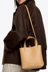 Bolso pequeño de piel vacuna color negro, con estructura rígida, doble asa de mano y correa larga ajustable para llevar al hombro o cruzado. Presenta cierre de botón a presión y logo metálico de la marca en el frente.