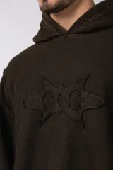 Buzo canguro de corte oversize, confeccionado en frisa de algodón con efecto de lavado gastado. Presenta un aplique decorativo cosido al corte en el centro del pecho, capucha amplia y bolsillo frontal tipo canguro.