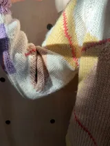 Cárdigan tejido de punto con diseño de rombos, en colores crema, violeta y detalles en rojo. Presenta cuello redondo, cierre frontal con botones de madera y textura acanalada en los bordes.