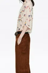 Camisa oversize de tejido fantasía color crema con estampado de pájaros y flores en tonos azules, amarillos, naranjas y rojos. Presenta finas rayas verticales en colores pastel y lentejuelas distribuidas por toda la prenda. Tiene cuello camisero, cierre frontal con botones y mangas cortas rectas con un ligero vuelo. Incluye un bolsillo de plastrón en el pecho izquierdo.
