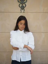 Camisa de manga larga confeccionada en broderie blanco, con diseño entallado, cuello alto y cierre frontal de botones. Presenta mangas abullonadas con hombros fruncidos y puños terminados en volados.