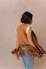Chaleco de gamuza sintética color tostado, con diseño abierto sin cierre. Presenta bordados decorativos en hilo blanco y flecos largos en el delantero y la espalda.