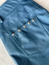 Pantalón de cuero ovino azul con textura de pitón. Presenta un bolsillo trasero decorado con cinco tachas metálicas doradas en forma de estrella. Tiene tajo externo en la pierna.