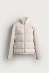 Cazadora acolchada de color beige con cuello subido, cierre frontal de cremallera y bolsillos de vivo en la cadera. Cuenta con mangas largas confeccionadas en tejido elástico y bajo acabado con elástico.